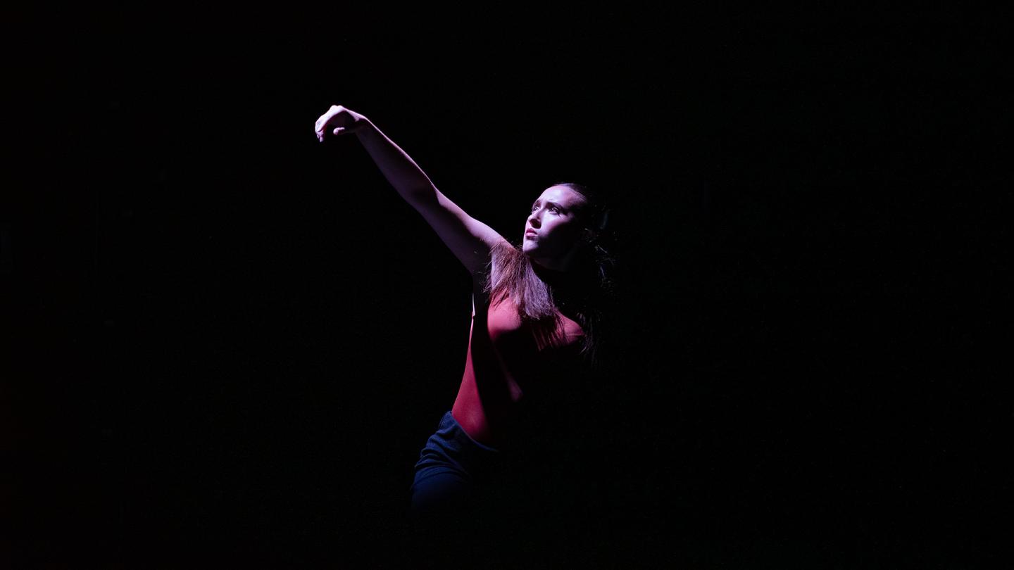 Woman against a black background wearing red, one arm raised, face lit and 