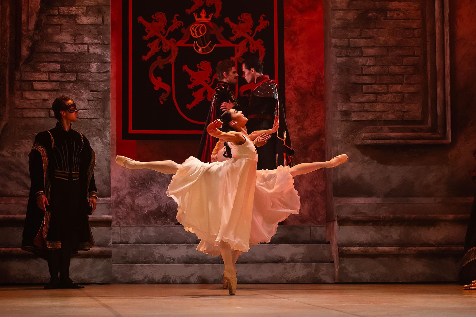 Dancer dressed in a white flowing dress dances elegantly, one leg raised behind her perpendicular to the floor while en pointe with the other
