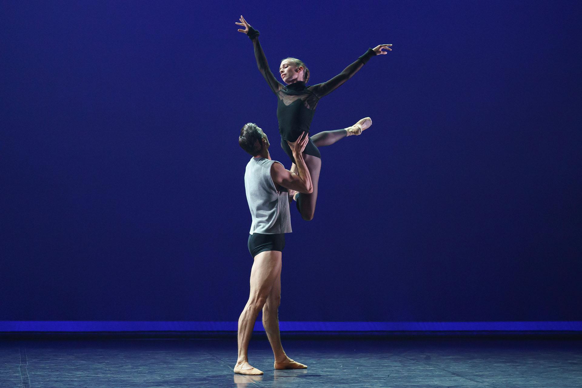 Dancer in a grey T-shirt and dark shorts lifts another wearing all black as she holds her arms wide and extends a leg behind her