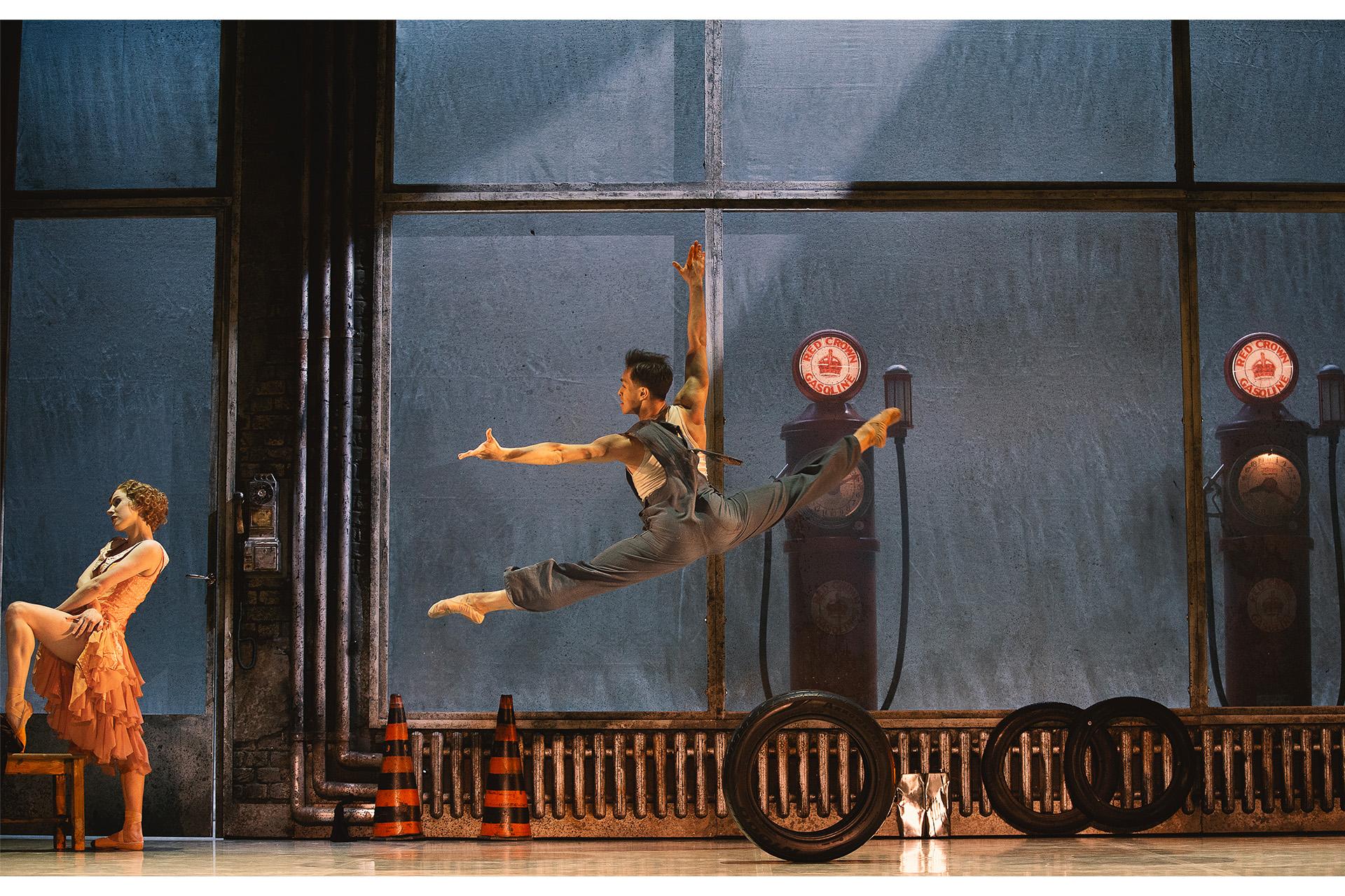 A male dancer jumping in splits on stage set like a car garage.